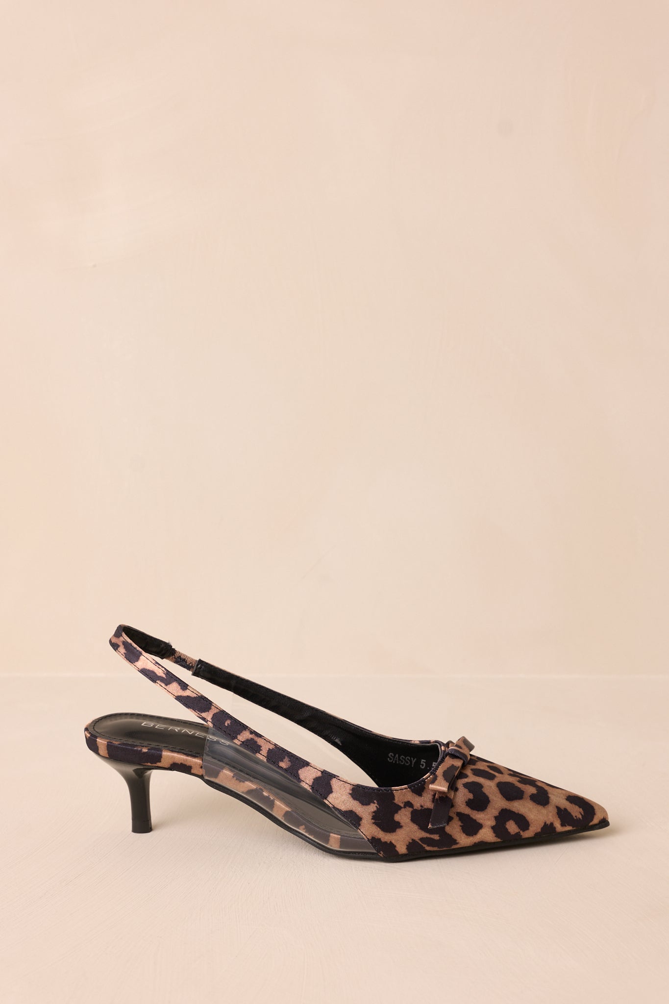 Side view of leopard kitten heels showcasing the pointed closed toe, delicate bow detailing, strap around the back of the foot, clear siding for extra support, and the short heel.
