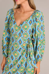 Close up view of this turquoise multi print dress that features a wide v-neckline, a self-tie waist feature, elastic shoulders, and elastic cuffed sleeves.