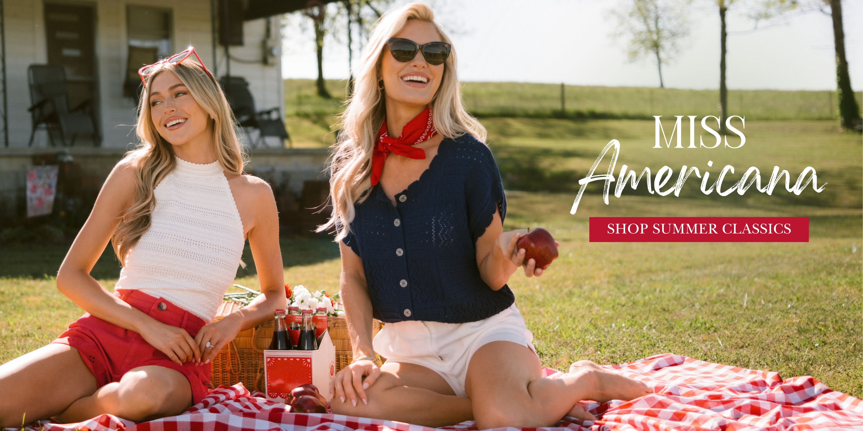Two women in red, white, and blue summer outfits sit on a picnic blanket outdoors, smiling with a basket of drinks and apples, styled in a “Miss Americana” theme and wearing outfits featured in Americana Collection.