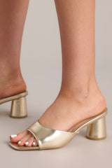 Close up view of these gold heels with square toe, slip-on design, strap over foot, short circular heel.
