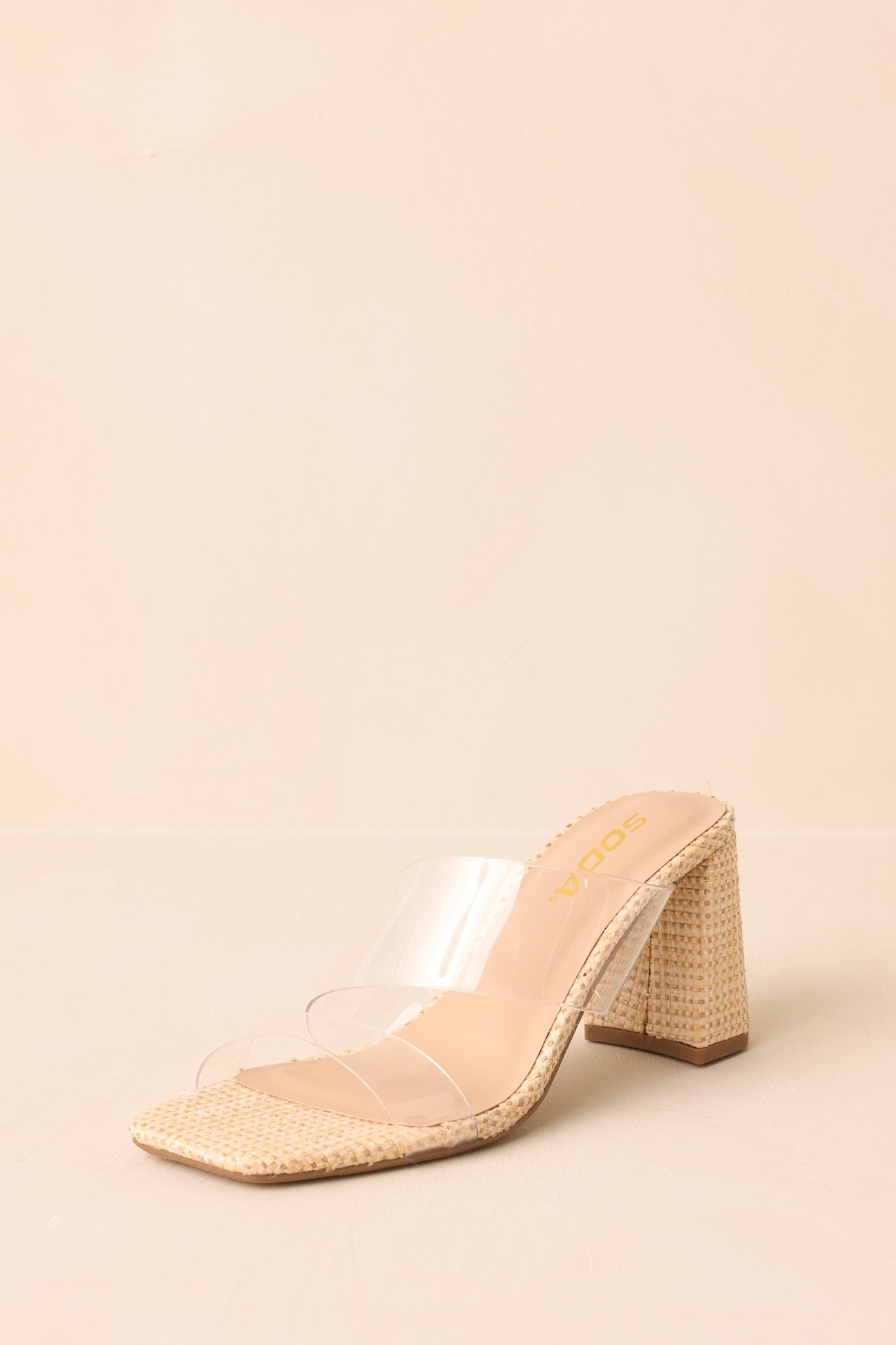 Natural raffia heels with clear straps displayed on beige backdrop, angled forward.