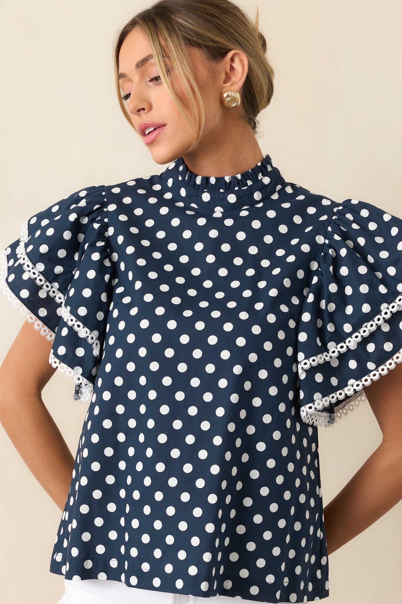 Navy and white polka dot top with a ruffle mock neckline and short ruffle sleeves, featuring ivory eyelet trim along the edges.