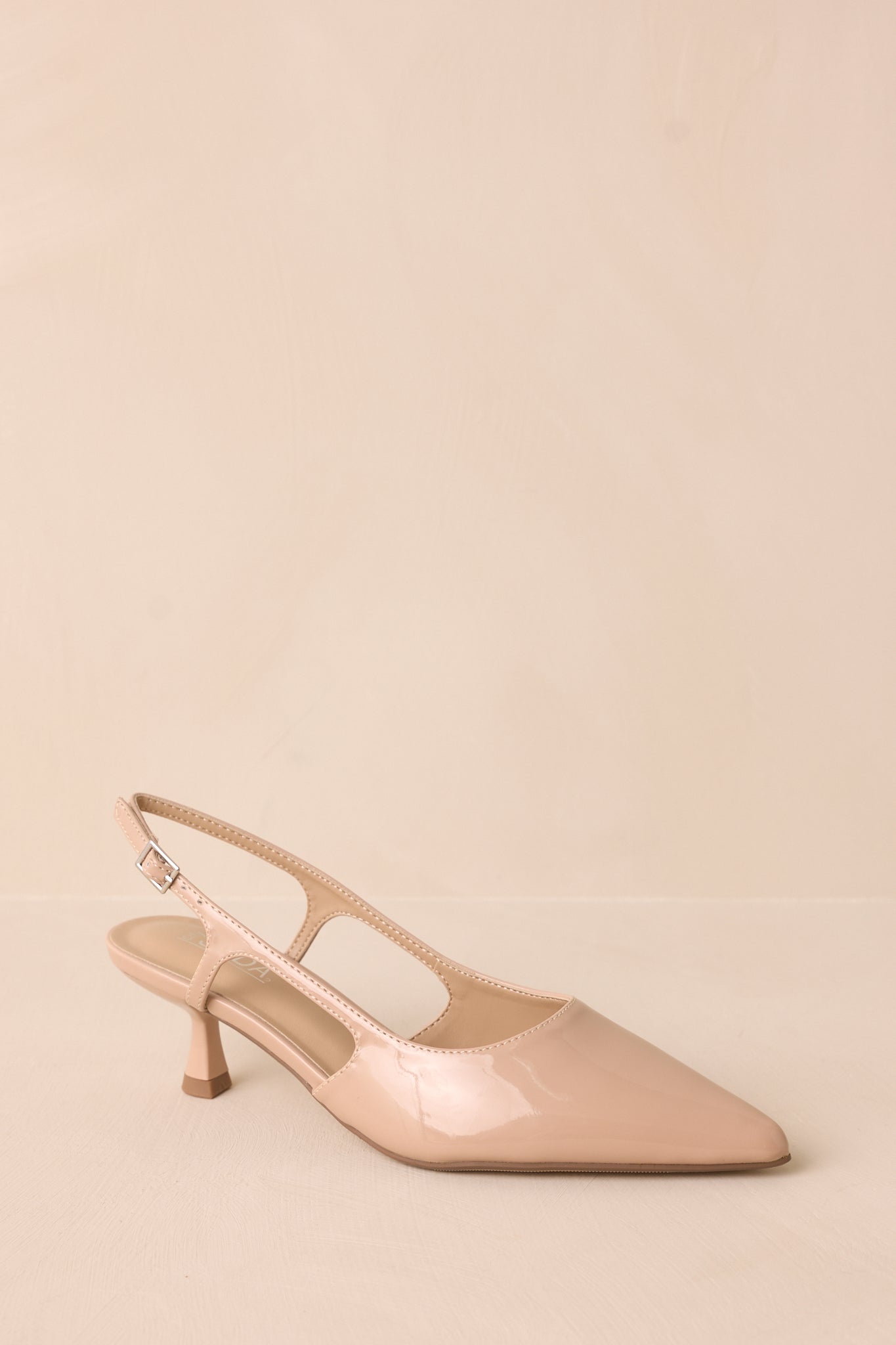 Nude patent pointed heel with gold buckle detail and side cutout, angled front view on backdrop.