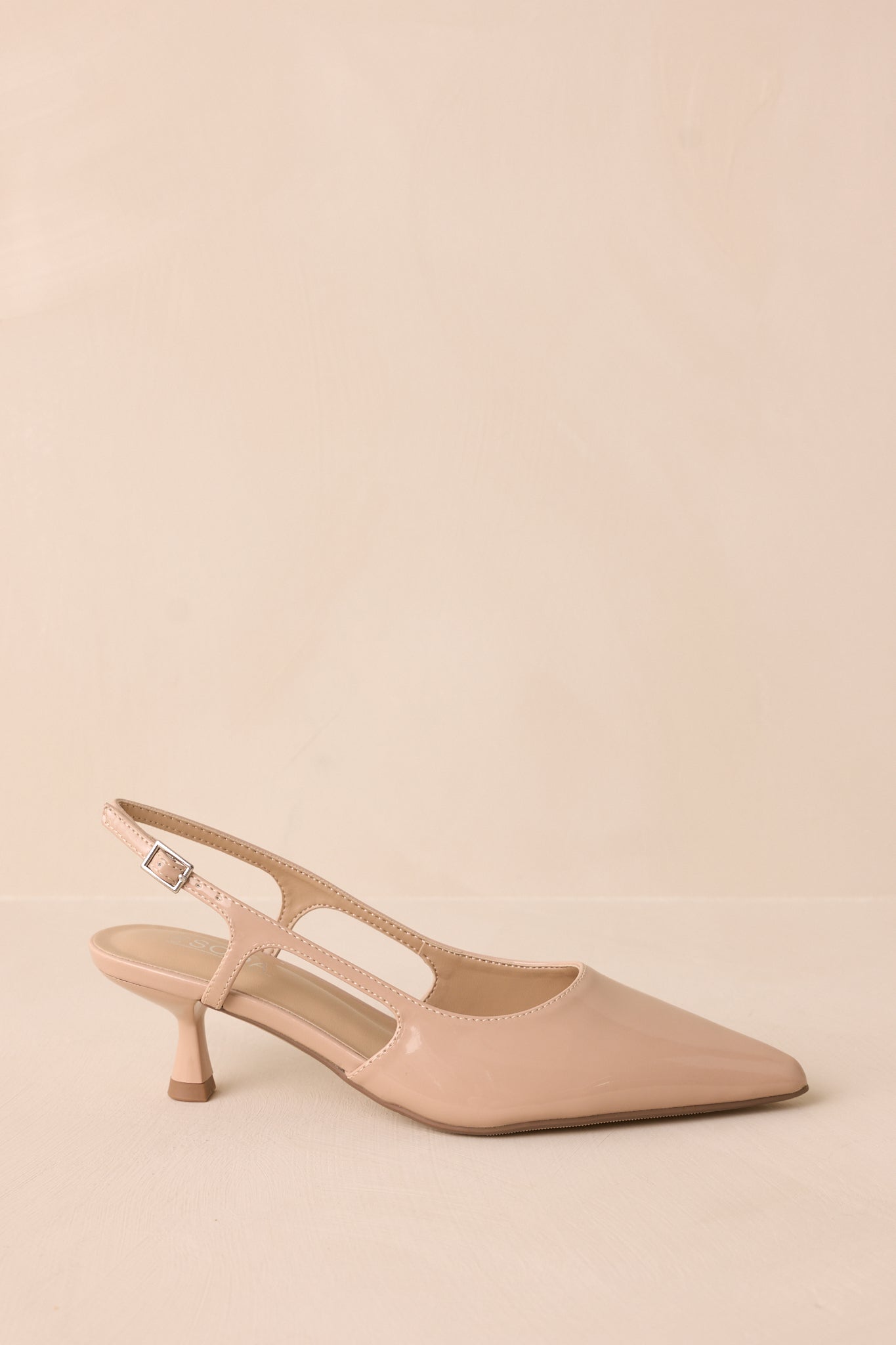 Nude patent pointed heel with gold buckle detail and side cutout, side view on backdrop.
