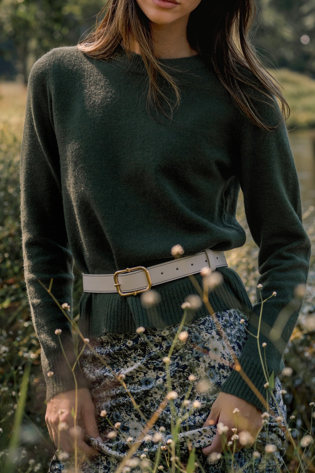 Close-up of an olive green knit sweater styled with a cream belt and blue floral skirt in an outdoor fall setting.
