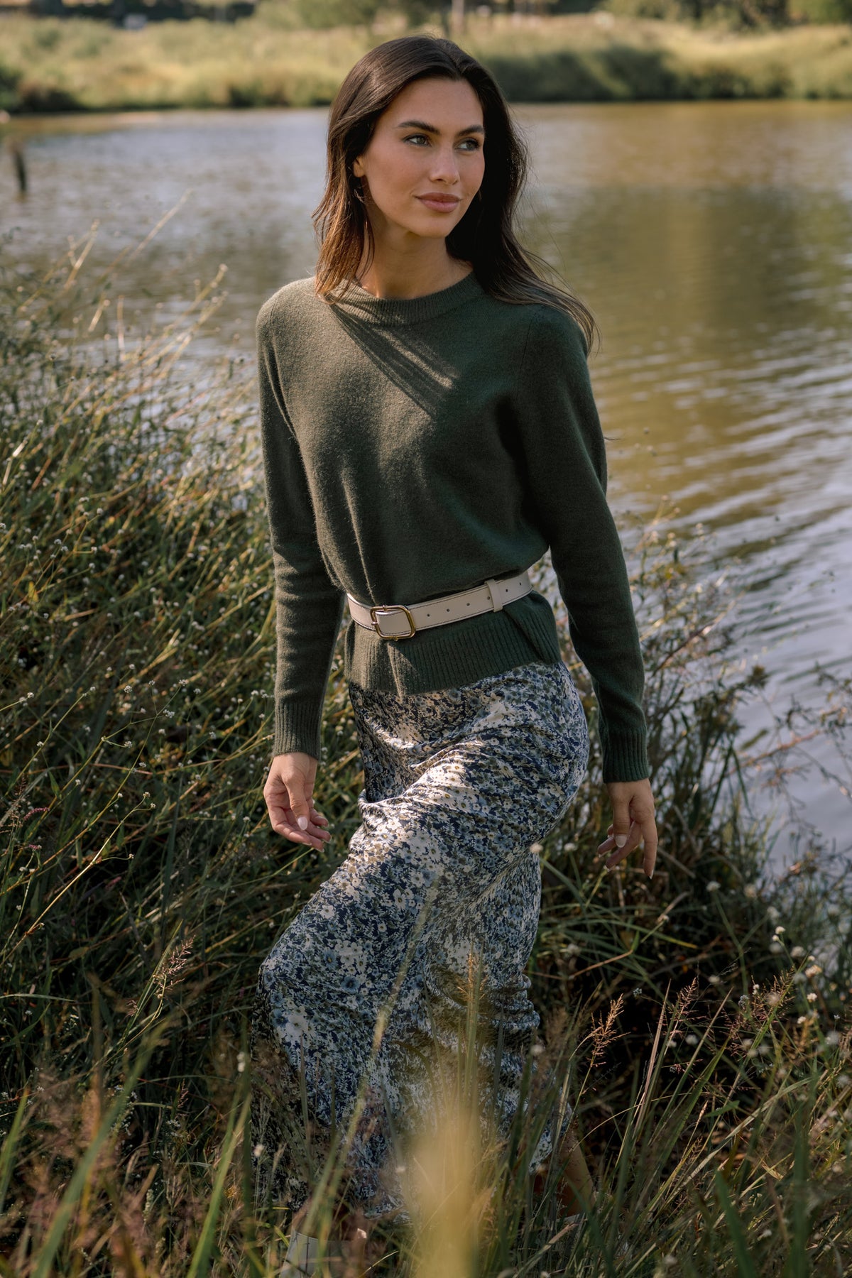 Woman wearing an olive green crewneck sweater, blue floral maxi dress, and cream belt beside a peaceful pond.