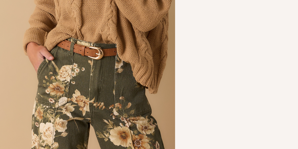 A person wearing a chunky beige cable-knit sweater, olive green floral pants with a brown belt, standing against a warm beige background.
