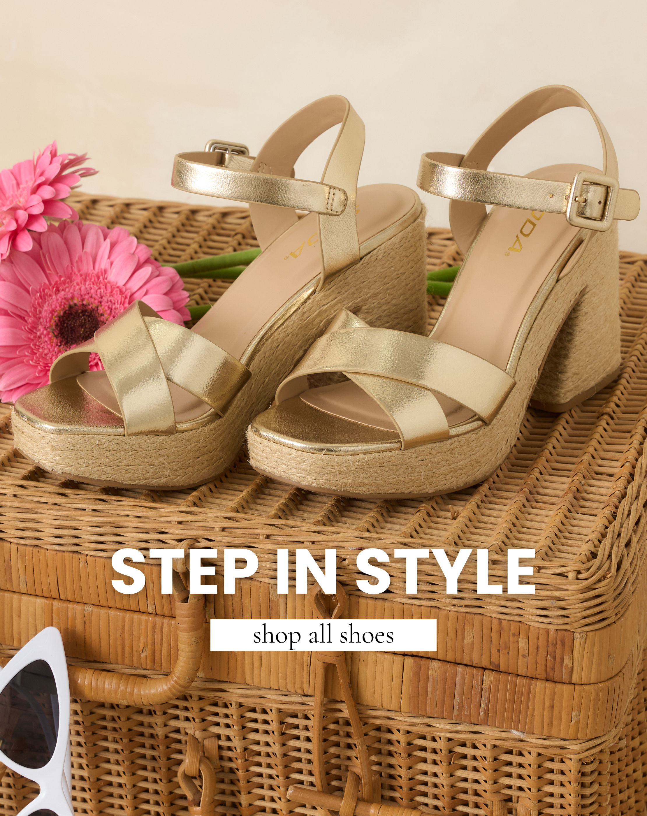 Gold platform espadrille sandals displayed on a woven wicker trunk with pink flowers for a chic spring shoe moment. This image leads to shop all shoes from Red Dress.