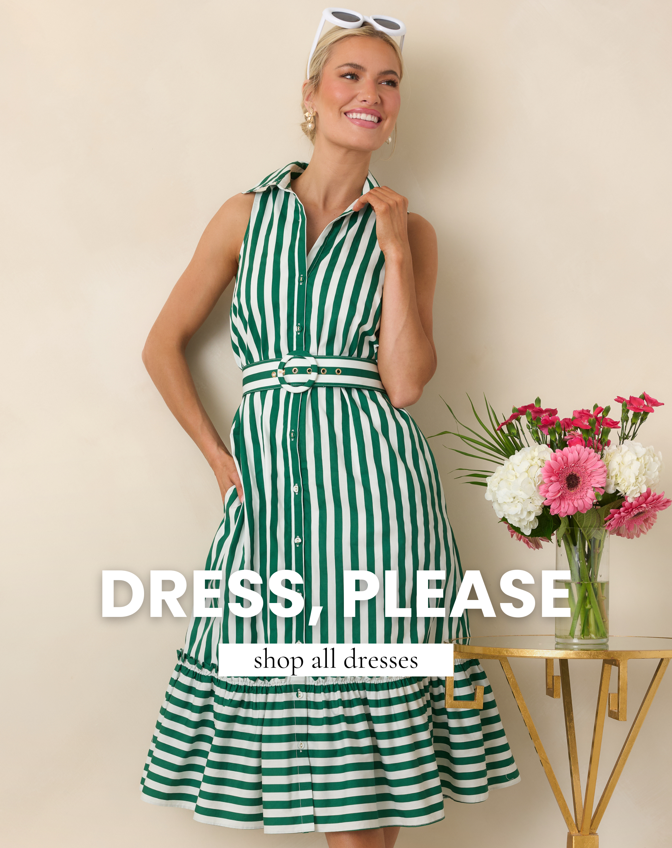 Woman wearing a green and white striped belted midi shirt dress styled with white sunglasses beside fresh pink and white flowers. This image leads to shop all dresses from Red Dress.