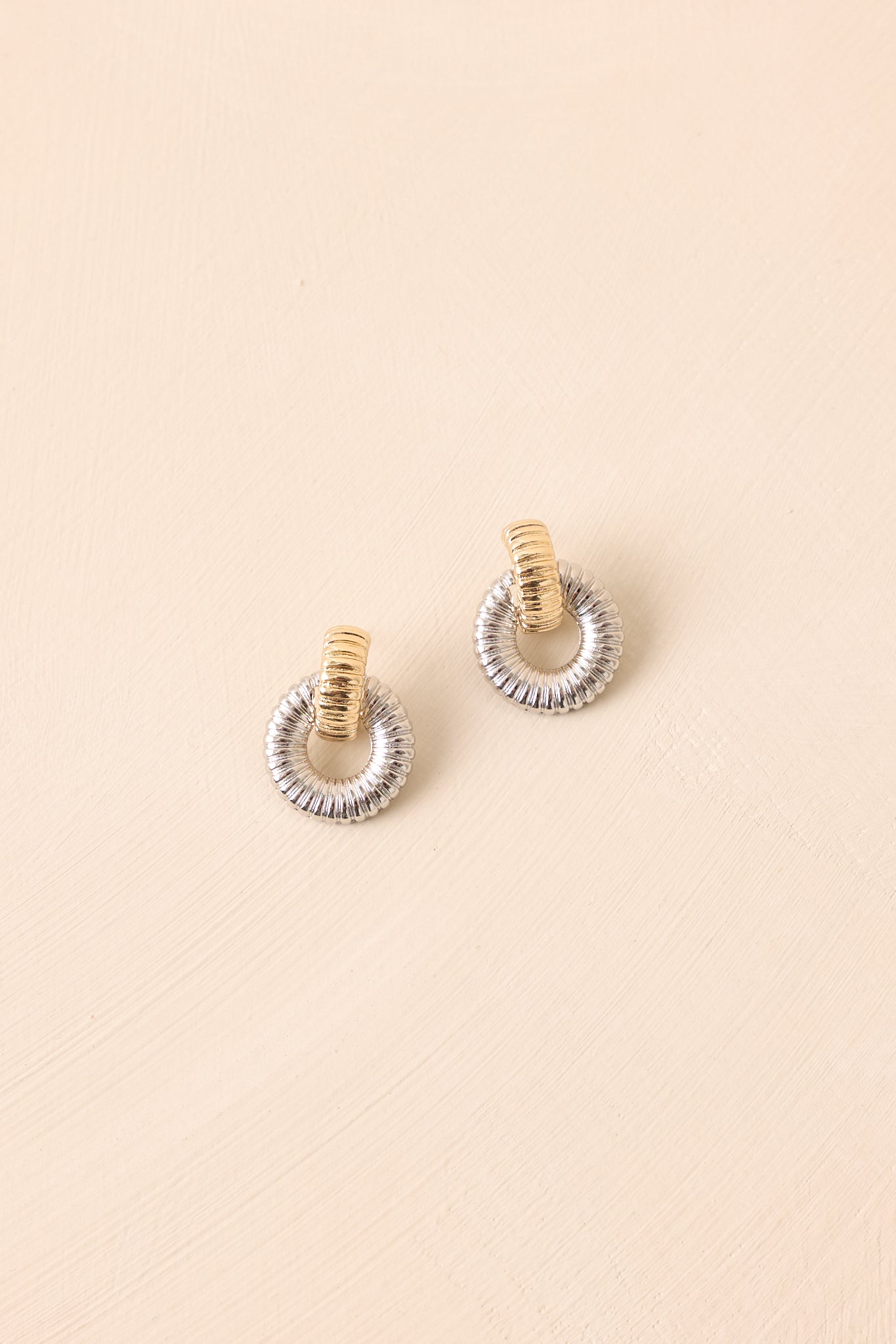 A pair of ribbed silver circular hoop earrings with a gold-tone center link displayed on a beige background.