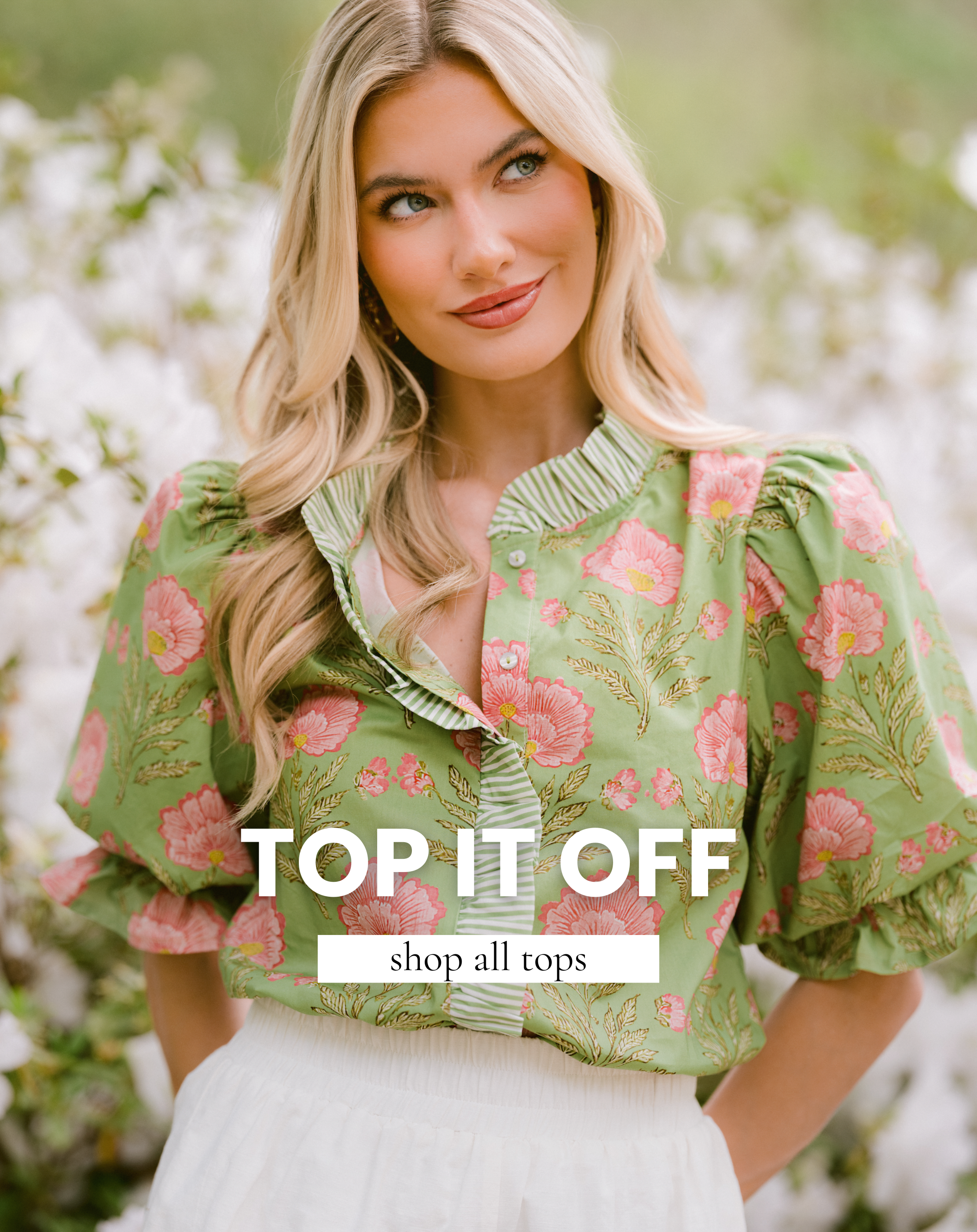 Blonde model wearing a green blouse with pink floral print and ruffled collar, styled with a white skirt, standing in a garden with blooming flowers—featured top available to shop.