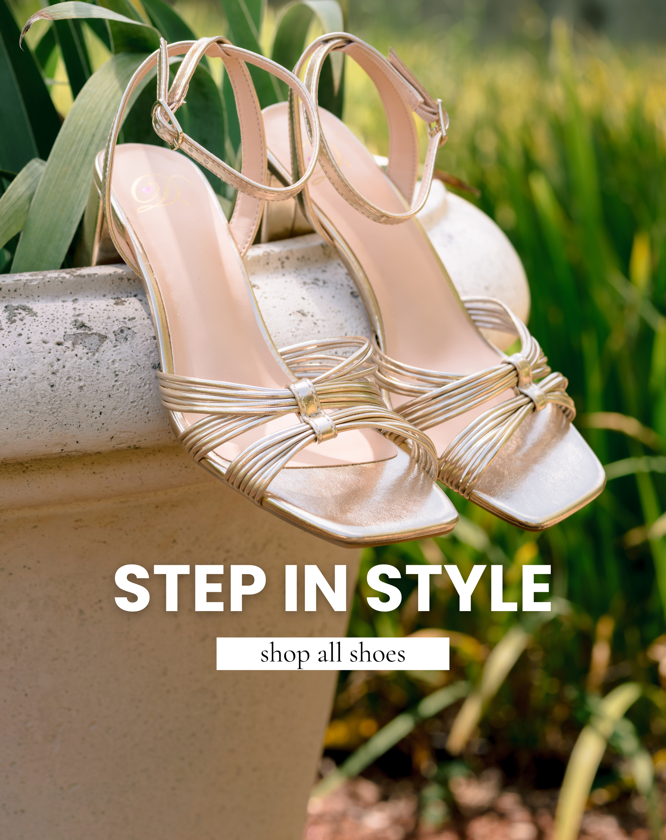 Pair of metallic gold strappy high heel sandals displayed on a stone planter with text promoting the shop all shoes collection.