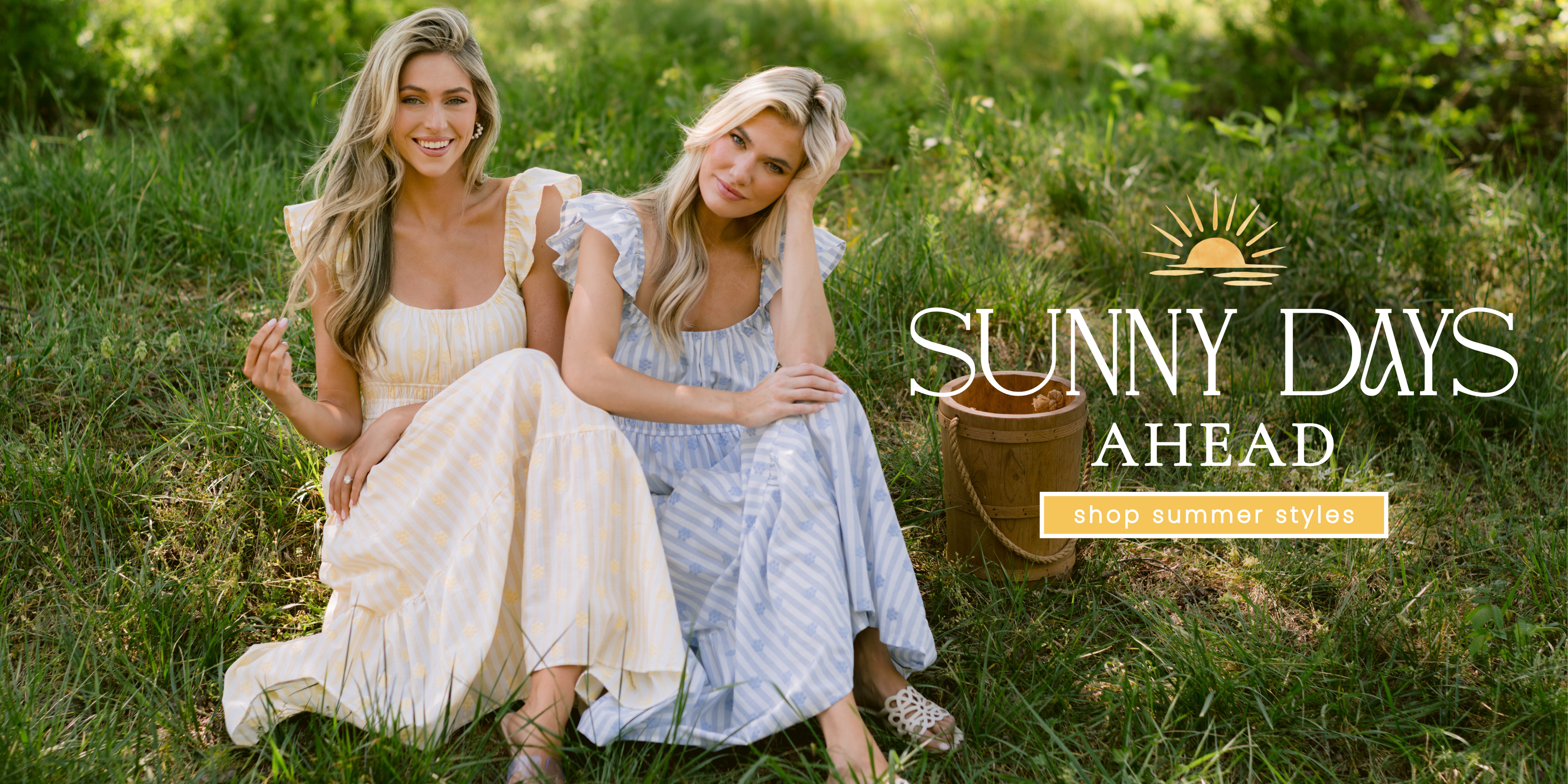Two models sitting in a sunlit grassy field wearing soft pastel striped maxi dresses with flutter sleeves in yellow and blue. Text overlay reads “Sunny Days Ahead” with a sun graphic above and a button-style banner that says “shop summer styles.”