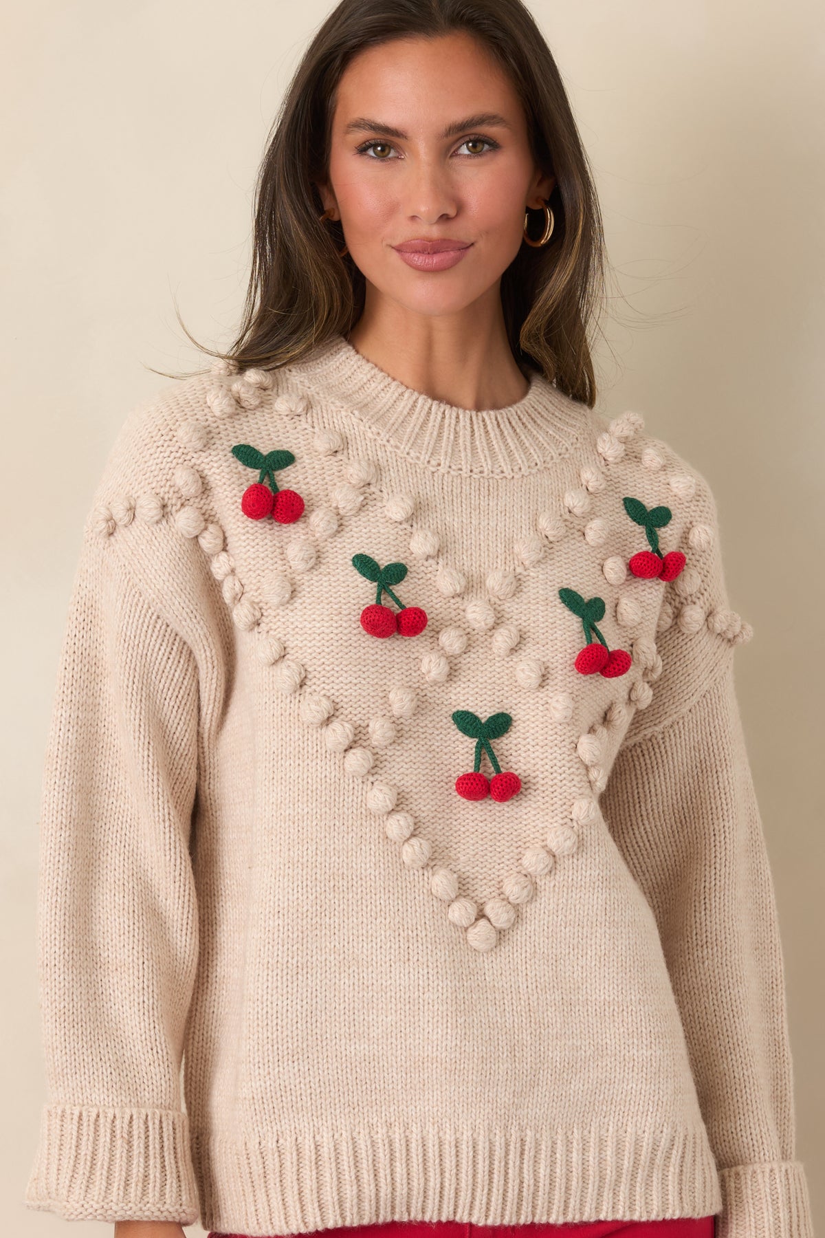 Angled front view of a beige cherry bobble knit sweater with dropped shoulders, long sleeves, and a ribbed hem.
