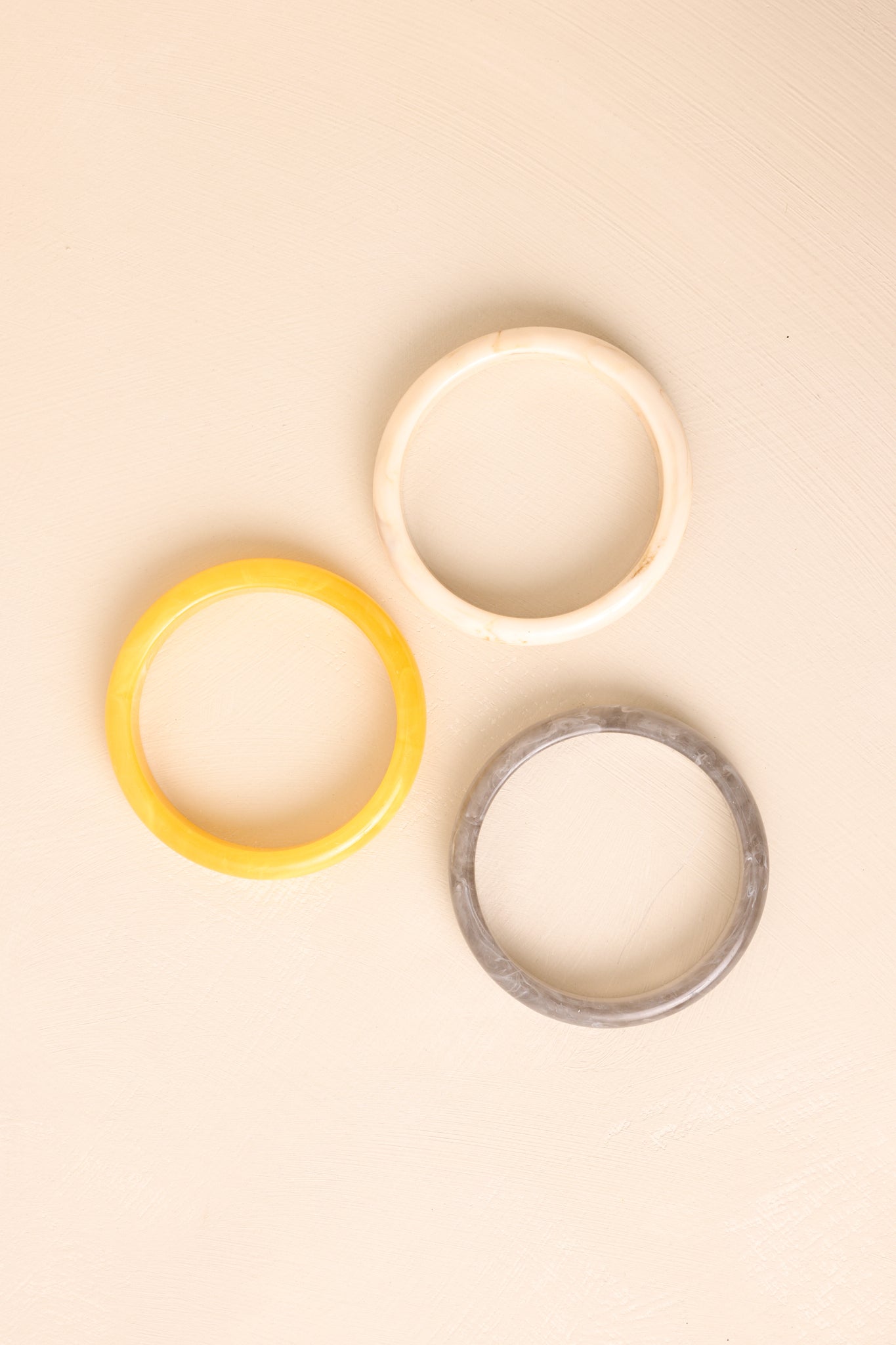 Set of three marbled bangles in soft yellow, ivory, and grey tones resting side by side on beige background.