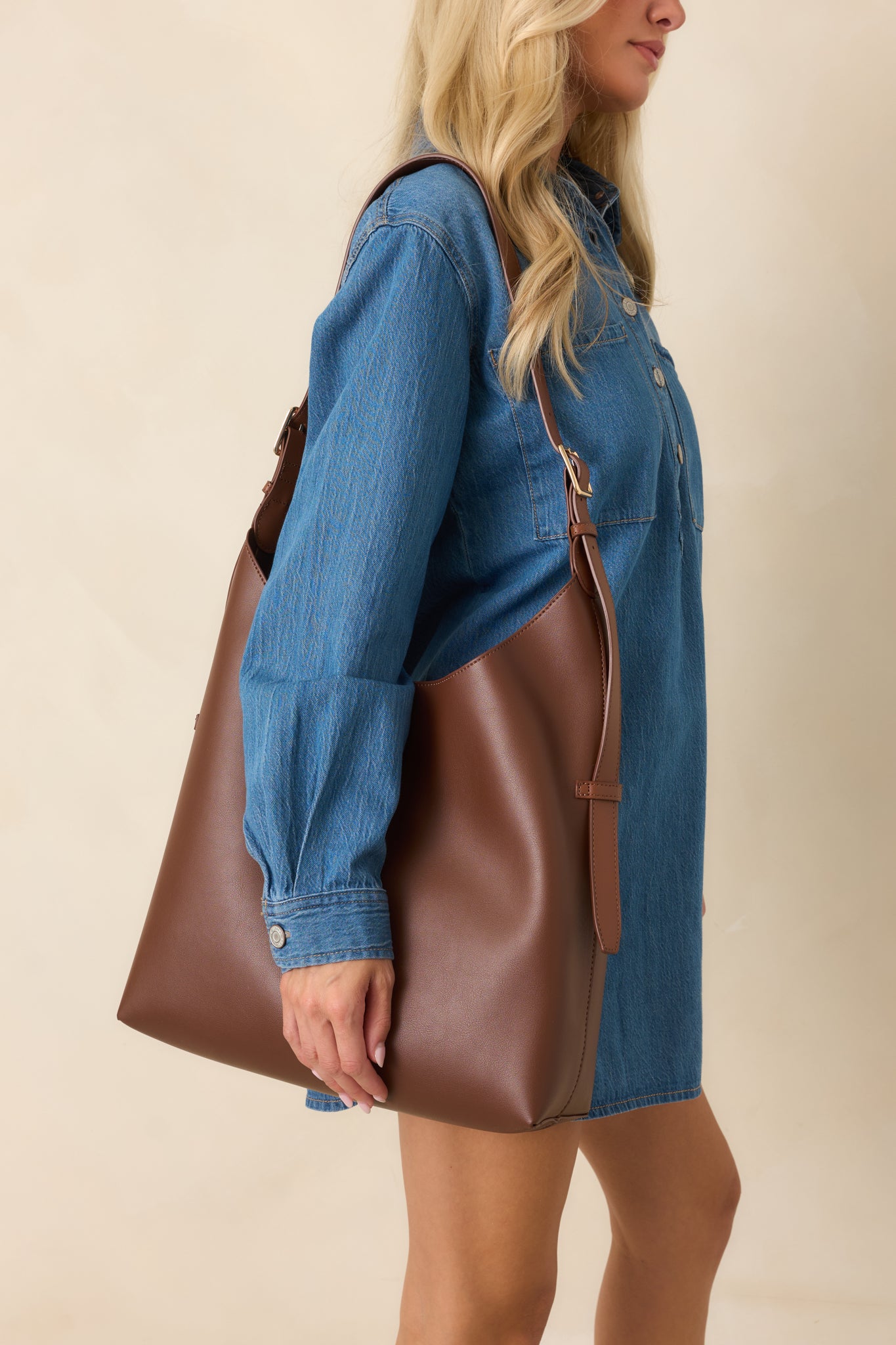 Women's chocolate brown faux leather hobo shoulder bag with an adjustable shoulder strap, styled with a blue denim shirt dress.