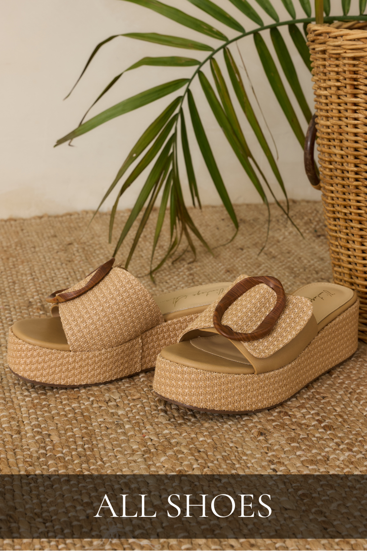 Raffia platform slide sandals with a wide faux leather strap and oversized buckle detail displayed on a beach rug, styled outdoors with palm trees in the background.