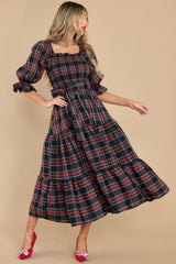 This dress has a mid-length, flared skirt reaching mid-calf and a fitted bodice with a square neckline. It's designed with puffed short sleeves and a smocked back for comfort. The fabric showcases a red and black tartan plaid pattern with white and blue lines. A removable sash belt emphasizes the waist. 