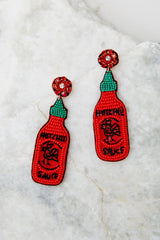Top view of these statement earrings that feature a beaded hot sauce bottle design, sewn details, felt back, and a secure lock back fastening.