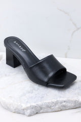 Close up view of these heels that feature a square toe, a thick strap across the top of the foot, a square heel, a faux-leather finish, and a slip-on design.