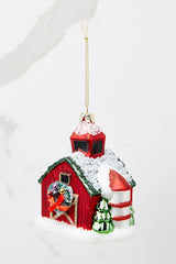 Angled front view of this ornament that features a barn shape with a green roof and a wreath on the door with gold accents.