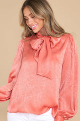 This coral colored top features a high neckline with an adjustable self tie, flowy balloon sleeves with two buttons at the cuff, and a relaxed fit.