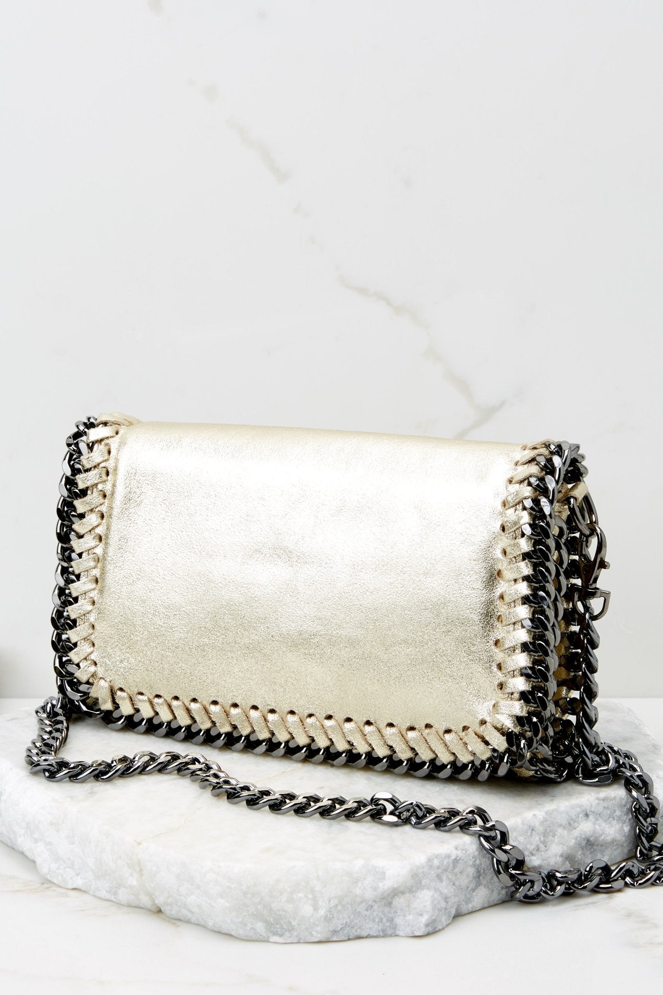 This gold clutch features a front flap that has a silver magnetic closure, an inside zipper, a removable leather strap, and wide decorative stitching around the edges. 