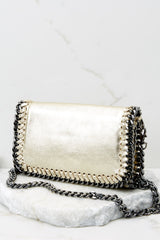 This gold clutch features a front flap that has a silver magnetic closure, an inside zipper, a removable leather strap, and wide decorative stitching around the edges. 