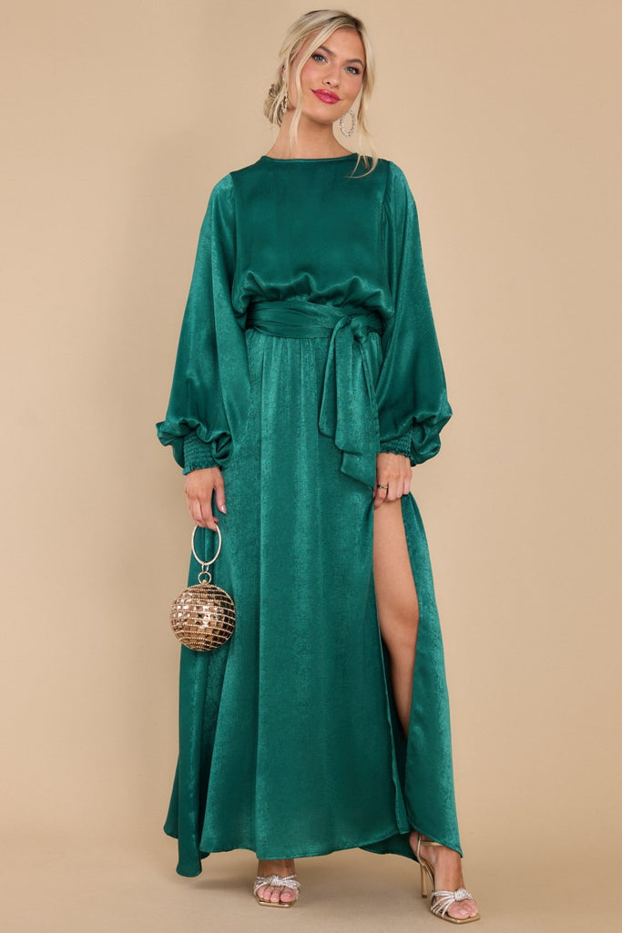 This dark green dress features a rounded neckline, keyhole cutout in the back with a button closure, balloon sleeves with smocked cuffs, an elastic waistband with a self-tie belt, and a shiny satin material.