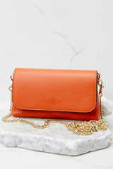 Front view of this orange crossbody bag that is made with real leather, has two detachable straps; one is a short matching leather strap, and the other is a curb chain crossbody strap, a magnetic snap closure, and measures 8