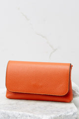 Close up view of this orange crossbody bag that is made with real leather, has two detachable straps; one is a short matching leather strap, and the other is a curb chain crossbody strap, a magnetic snap closure, and measures 8