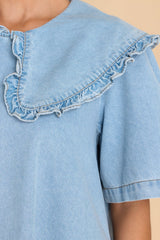 Close up view of this dress that features a high neckline with a ruffled collar, short sleeves functional zipper in the back, pockets, and chambray design.