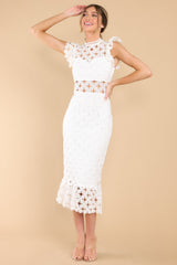 Full body view of this crochet lace midi dress that features a high neckline with an illusion sweetheart neckline, ruffled shoulders, a peek-a-boo waistline which gives way into a sheath silhouette fit, and a flounced hem. Dress has a visible back zipper.