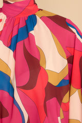 Close up view of this top that features a high neckline, an adjustable tie around the neck, and abstract pattern in shades of red, orange, yellow, blue, ivory, and pink.