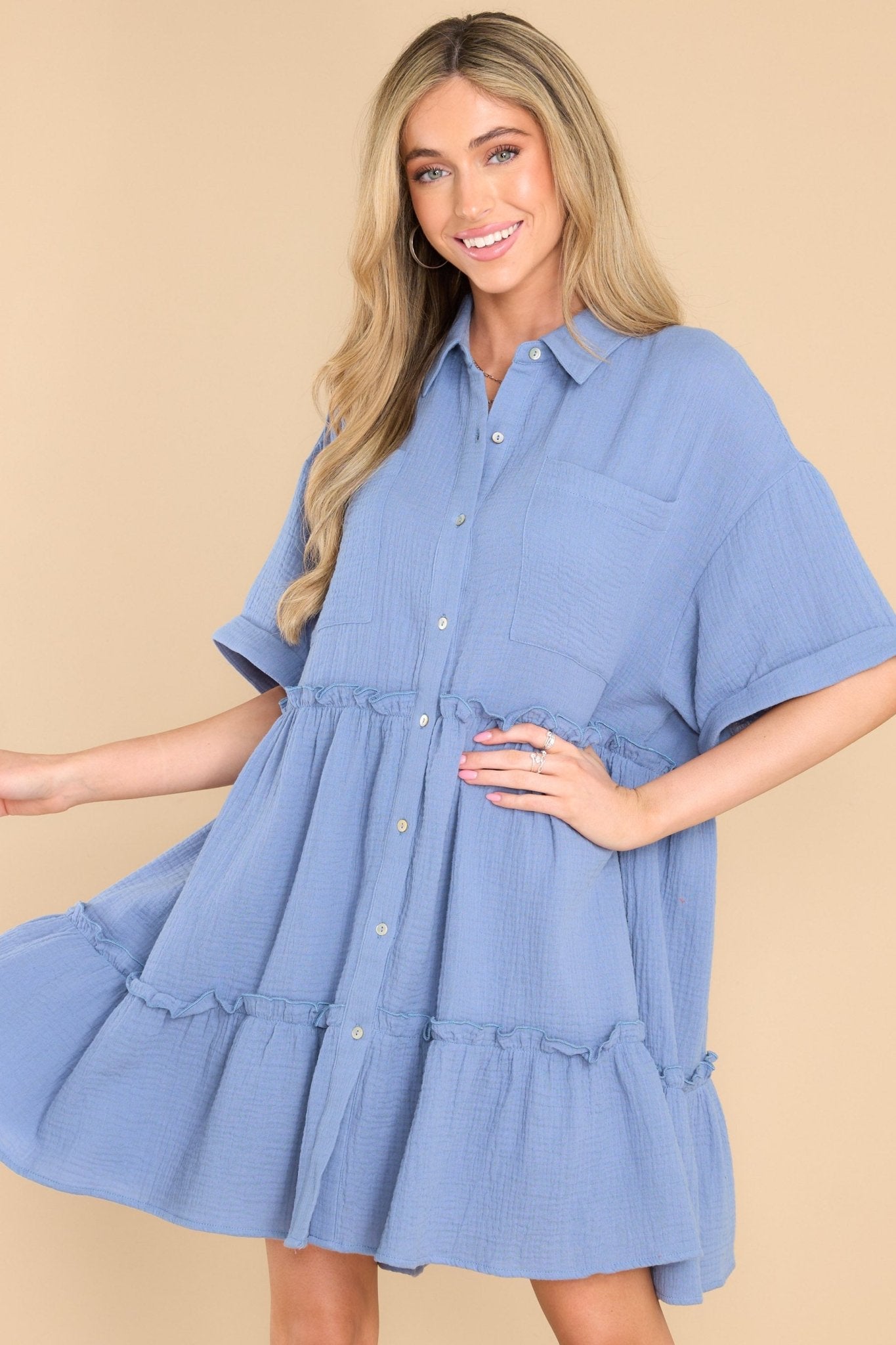 This blue dress features a collared neckline, functional buttons down the front, functional pockets at the bust, short sleeves with folded cuffs, and ruffle detailing throughout the skirt.