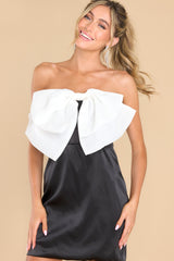 Front view of this dress that features a strapless neckline with a non-slip rubber strip, a zipper in the back, a large bow detail at the center of the bust, boning in the bust section, and a satin feel. 