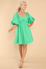 This green dress features a square neckline, puff sleeves with elastic cuffs, adjustable self tie over an open back, pleated detail over bust, and flowy skirt.