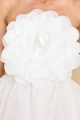 Close up view of this dress that features a strapless neckline, a rubber non-slip band around the top of the bust, a smocked section and functional zipper in the back of the bust, and a three-dimensional flower detail on the front of the bust.